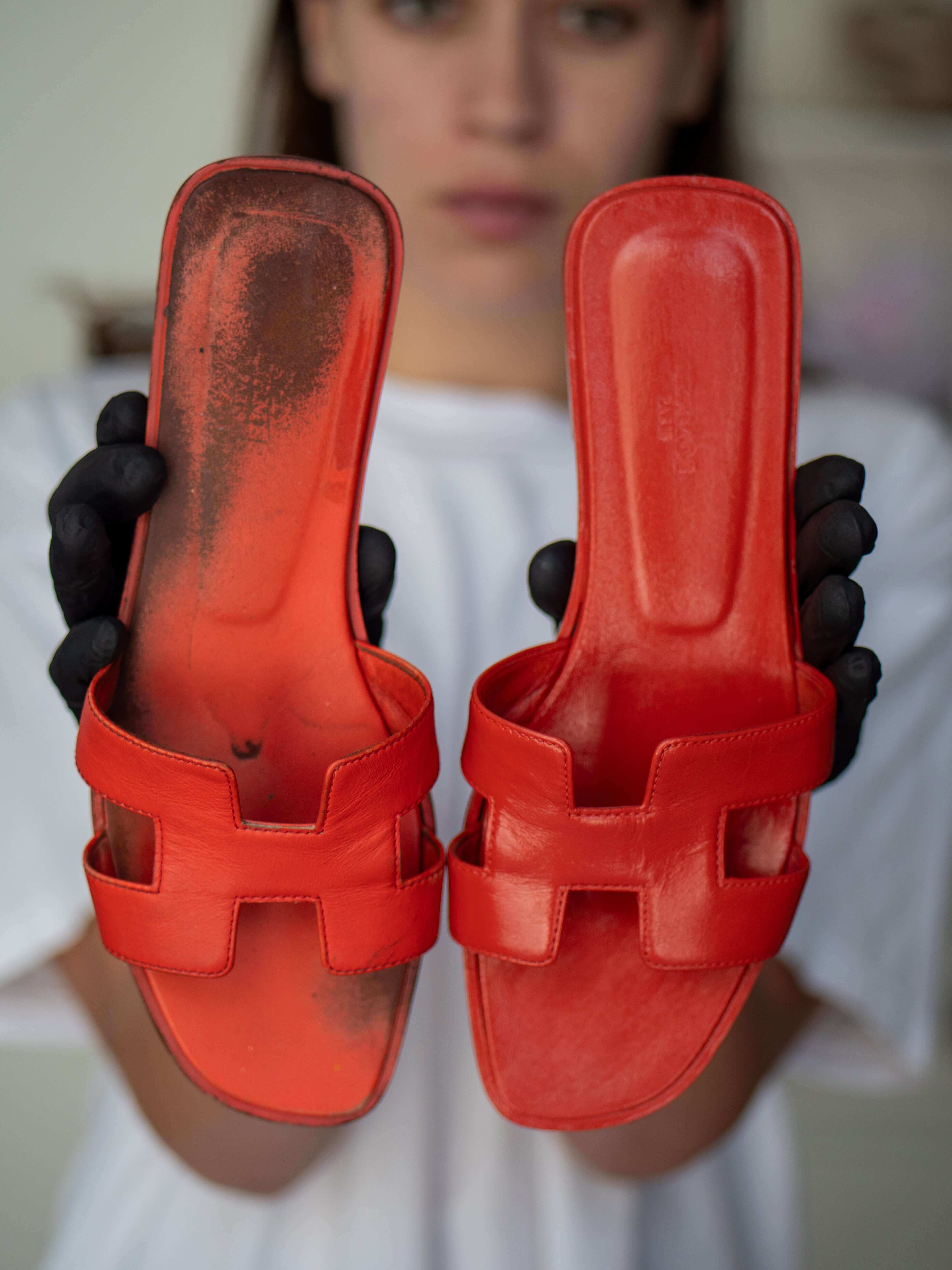 Before and after restoration of Hermès Oran sandals showing heavy wear on left pair and fully cleaned, refreshed leather on right pair, held by artisan in gloves