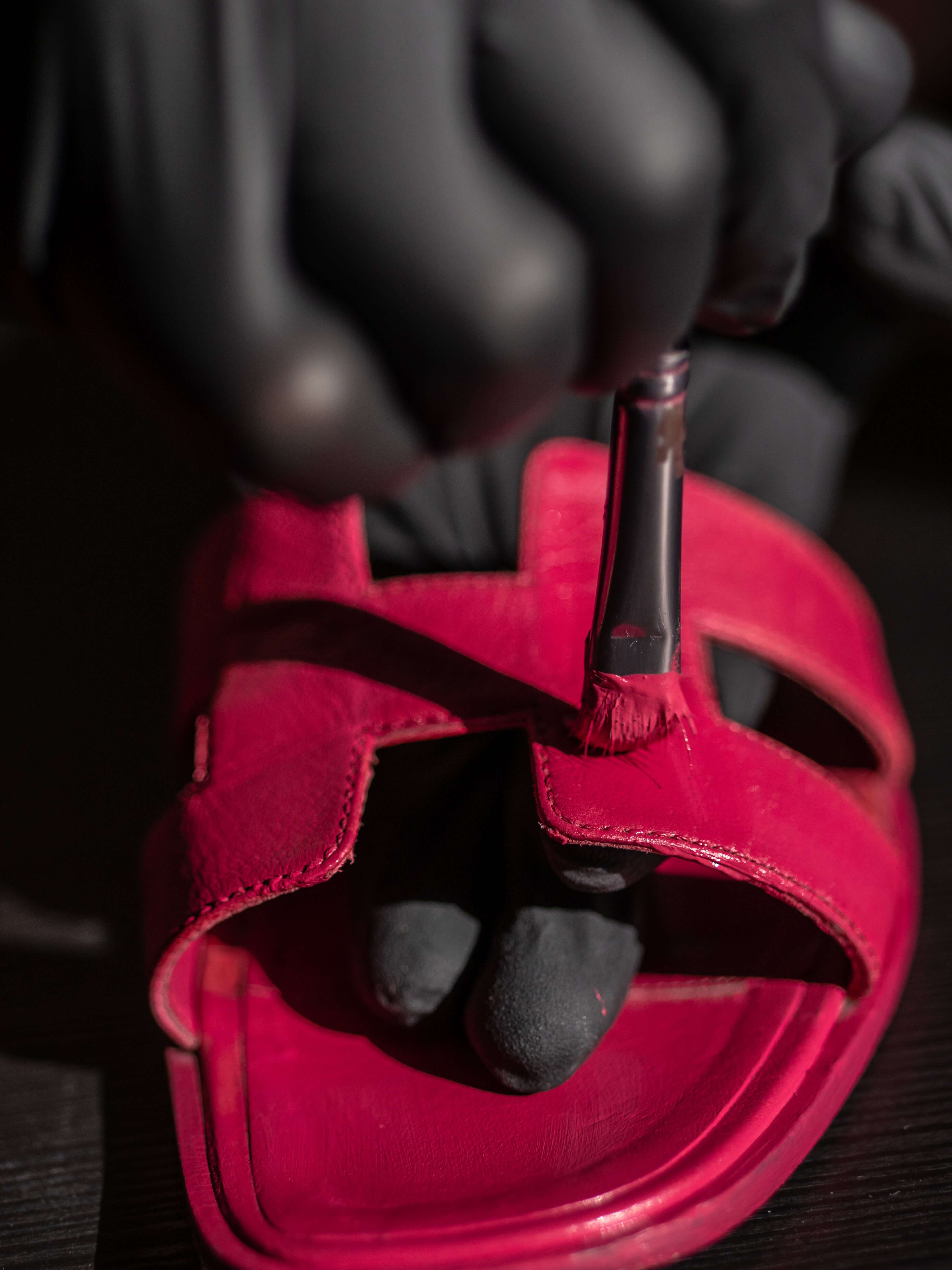 Artisan hand-dyeing a red Hermès Oran sandal using a precision brush during professional leather color restoration service
