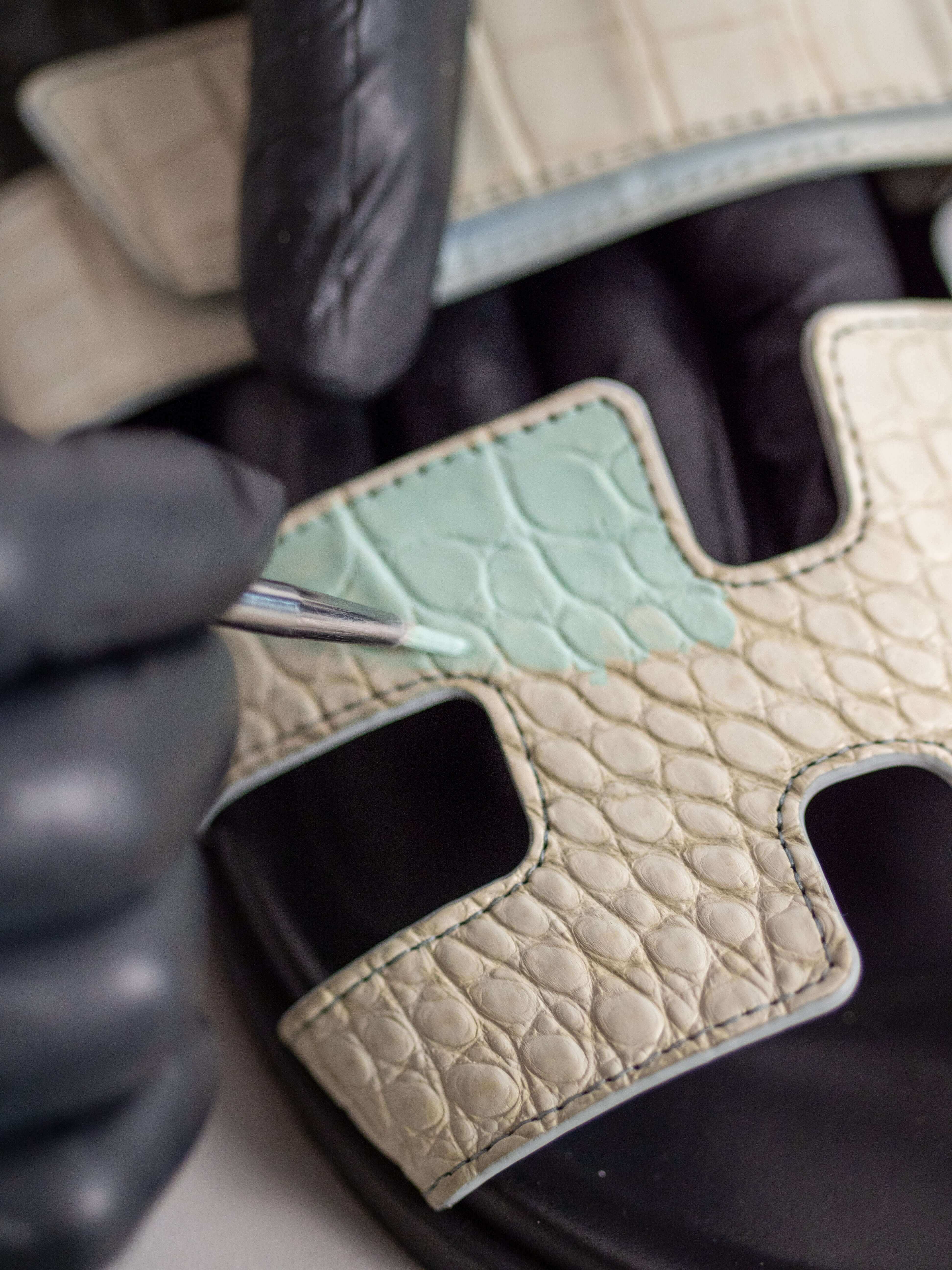 Hermès Chypre sandal in exotic leather being hand-painted to restore original color after fading, with artisan using precision brush for color matching